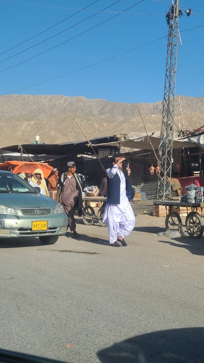 The image captures a street scene in Bostan, Pakistan. A group of men are gathered around a light green Toyota Corolla with license plate AOT-226. Several of the men are in traditional Pakistani clothing. One man is lying on a cart, while another is standing near the car. Overhead are power lines and towers. A mountain range forms the backdrop to the scene.