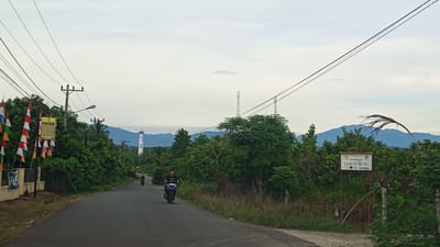 The image depicts a rural roadside scene in Kilometer Delapan, Indonesia. A paved road curves through a landscape dominated by lush green vegetation and trees. In the foreground, a man rides a motorcycle on the road. Two other motorcycles are visible in the distance, also on the road.
On the left side of the road, there is a wall with several flags hanging from poles, and a sign that reads "POLSEK". A utility pole with wires runs along the roadside. In the background, a white lighthouse or tower stands tall amidst the trees, with mountains visible behind it. Two cell towers are also present in the distance. On the right side of the road, a sign with various symbols and text is mounted on a red frame. The sky is overcast with a pale, cloudy atmosphere. The overall impression is of a quiet, somewhat remote location.