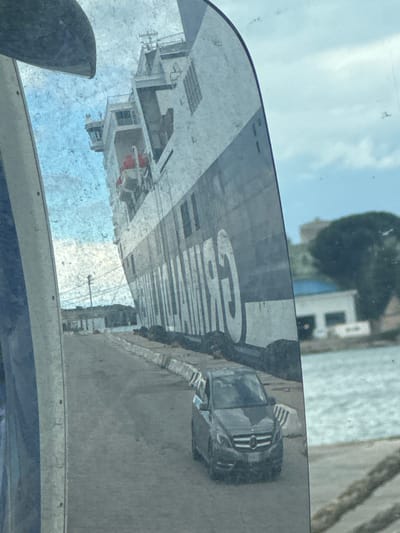 The image presents a reflection captured in a dirty vehicle side mirror, depicting a port scene in Brindisi, Italy. A large, multi-deck passenger and vehicle ferry, identifiable by the reversed "GRIMALDI" text on its hull, dominates the left and central portions of the reflection. The vessel is primarily white with a dark horizontal stripe, featuring visible windows, the bridge structure, and at least one red lifeboat. The ship appears to be docked at a concrete pier.
On the paved pier, in the foreground of the reflection, a dark grey Mercedes-Benz B-Class vehicle is positioned, facing away from the observer. The pier's edge is delineated by light-colored concrete barriers, some showing signs of wear and staining. Large, dark, cylindrical objects, possibly fenders or bollards, are situated along the pier near the ship's hull. In the mid-ground, a utility pole is visible.
To the right of the paved pier, a body of water, likely a harbor or sea, is visible. Across the water, faint structures include a building with a blue roof and distant green foliage. The sky above is partly cloudy. The mirror's surface is notably smudged and dirty, introducing a speckled, hazy texture to the entire reflected image. No identifiable individuals are visible within the scene.