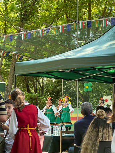 An outdoor cultural event is depicted in Bolyarovo, Bulgaria, under a large green canopy tent, with lush green trees forming the background. A string of small pennant flags, alternating between the Bulgarian flag (white, green, red horizontal stripes) and the European Union flag (blue with yellow stars), is strung above the canopy. On a stage beneath the tent, three young women or girls are performing, dressed in traditional Bulgarian folk costumes featuring white blouses, green dresses with elaborate trim, and colorful floral headpieces. Each performer stands before a microphone. Water bottles are arranged on a table at the front of the stage. Behind the performers, a backdrop with horizontal green, yellow, and red stripes is visible, along with two flower-shaped balloon decorations (one green with a yellow center, another white with a red center). In the foreground, several spectators are seated, facing the stage. A person with a ponytail, wearing a red sleeveless garment over a white blouse with a yellow tie-belt, is seen from behind, accompanied by a young boy. Further back, a man in a dark blue jacket and another person with long, wavy hair are also observed from behind, attentively watching the performance.