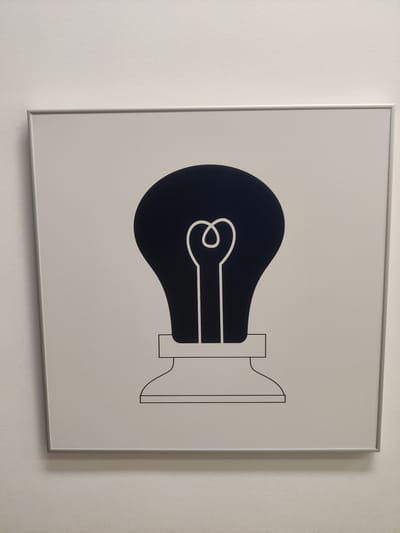 This is a photograph of a framed, minimalist illustration of a lightbulb. The illustration is in black and white, with a dark blue or black lightbulb silhouette and a white filament inside. The lightbulb is depicted in its base, with a simple, stylized stand. The artwork is framed by a thin, metallic silver frame and is mounted on a textured white wall. The lighting suggests an indoor setting, likely a well-lit room or hallway. No people are visible in the image. The overall impression is one of simplicity and modern design. The location context of Thessaloniki, Greece, does not offer any specific visual cues within the image itself. There is no visible text in the image.