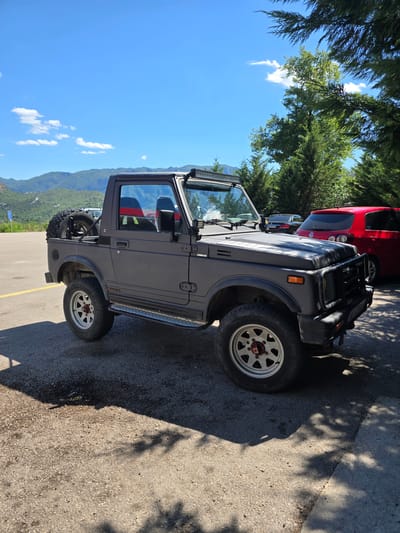 A matte gray, convertible Suzuki Samurai JX is parked on a paved surface in a lot, with a red hatchback and a dark sedan visible behind it. The Samurai has a spare tire mounted on the rear, a light bar across the top of the windshield, and aftermarket wheels. The scene appears to be in a mountainous, semi-rural area, with lush green trees and a clear blue sky overhead. A road sign is visible in the background to the left. The parking lot has yellow painted lines. The ground shows some wet patches and shadows cast by trees. The location is identified as Sotonići, Montenegro.