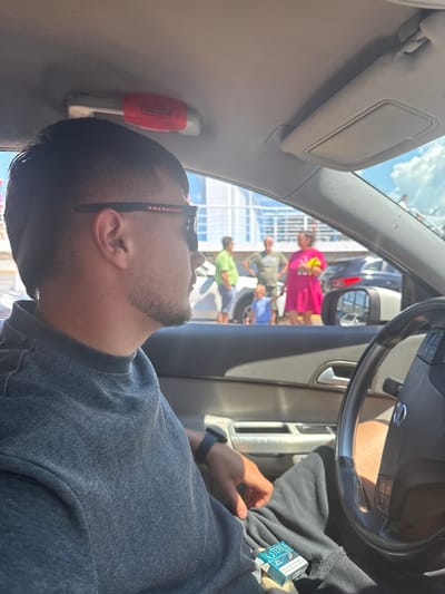 A medium shot captures a man from the chest up, seated in the driver's seat of a car. He is wearing a dark gray t-shirt and dark pants. His left hand rests on his lap, near a pack of cigarettes. His right arm is not visible. The man has dark hair and is wearing black-rimmed glasses with "PRADA" printed on them. He is looking to his right, out the window.
Through the car's windows, a blurry outdoor scene is visible. Several people, including two men, a woman in a bright pink dress, and a young child in a blue shirt, are standing outside. They appear to be near a building or structure with a fence or railing. The sky is blue with some clouds, suggesting it is daytime. The context of Kavala, Greece, is not directly evident from the visual information in the image.