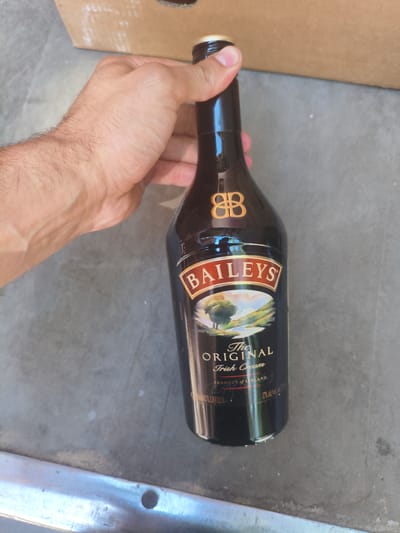 A person's arm and hand are visible, holding a dark brown glass bottle of Baileys Irish Cream liqueur. The bottle has a gold logo with the letters "BB" and a label with "BAILEY'S THE ORIGINAL Irish Cream PRODUCT OF IRELAND 70cl/0.7L LIQUEUR 17% alc/vol". The bottle is positioned vertically on a grey, textured surface, possibly concrete. In the background, a portion of a cardboard box is visible. The lighting suggests an outdoor or well-lit indoor environment. The image does not contain any visual information that would allow for the determination of its location in Alchevsk, Ukraine, or any other specific geographic area.