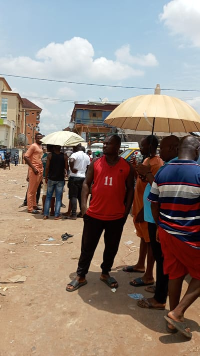 This image captures a bustling street scene in Ogbunike, Nigeria, under bright daylight. A group of approximately 10-15 men are gathered on an unpaved, dusty ground littered with debris. The sky above is clear with scattered white cumulus clouds.
In the foreground, a man in a red sleeveless shirt with the number "11" printed on it and black pants, wearing black slides, stands facing slightly to the right, looking into the distance. To his right, under a large, light-colored open umbrella, several men are congregated. One man wears an orange tunic and another a blue and red striped t-shirt. Another man under this umbrella appears to be holding a small red object. Further to the left, another large, light-colored umbrella provides shade for a smaller group of men, one wearing a black sleeveless shirt and blue jeans, and another in a white t-shirt. On the far left, a man in a peach-colored traditional outfit (tunic and trousers) stands, also wearing sandals.
The men appear to be casually interacting, standing, and observing their surroundings. Their postures suggest a relaxed gathering or waiting. Partially visible vehicles, including what looks like a white bus or van and a yellow commercial vehicle, are in the background. The background also features multi-story buildings, some with satellite dishes, indicating an urban or semi-urban environment. The strong shadows suggest it is midday or early afternoon, requiring the use of umbrellas for sun protection. The overall atmosphere is casual and typical of a local street gathering.