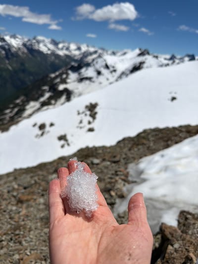 A close-up shot shows a person's hand holding a clump of snow. The hand is lightly tanned with some visible veins. The snow is white and crystalline, appearing to be melting slightly. In the background, a snow-covered mountain slope is visible, with patches of dark rock. Above the mountain, a bright blue sky with fluffy white clouds suggests a sunny day. The overall scene conveys a sense of being in a cold, mountainous environment, possibly during a hike or an outing in nature. The lighting suggests it is daytime, likely late morning or afternoon due to the brightness of the sky. The location is identified as Верхне-Тебердинское сельское поселение, Russia.