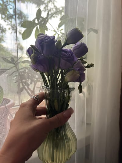 In the city of Zaşağan, Kazakhstan, a hand with a silver ring on its index finger is holding a green glass vase filled with a bouquet of purple flowers. The vase is positioned in front of a sheer white curtain, which is partially drawn back to reveal the silhouette of tree branches and leaves against a bright sky. The flowers, primarily in shades of purple with some lighter buds, are arranged in the vase, with their stems visible through the glass. The lighting suggests it is daytime, with natural light filtering through the window and the curtain. The image captures a still-life composition of flowers and a hand against a window background.