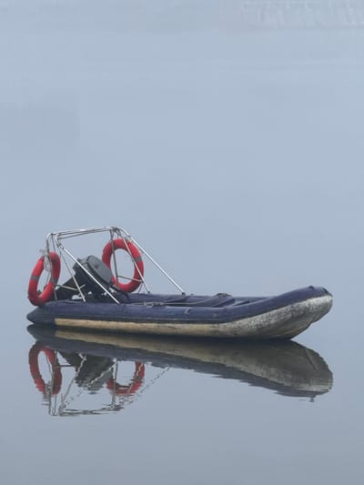 A blue and gray inflatable boat with a metal frame and two red life preservers is floating on calm, gray water. The boat has an outboard motor mounted on the back. The sky is overcast and the overall scene is muted and hazy. The boat is positioned diagonally in the frame, with the bow pointing towards the right. The reflection of the boat is visible on the water's surface. The image is devoid of any visible human presence or any specific landmarks that would indicate its location within Da Lat, Vietnam. The environment appears to be a body of water, possibly a lake or a quiet bay, under a diffused light.