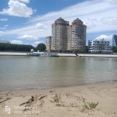 The image depicts a waterfront scene in the city of Atyrau, Kazakhstan, likely along the Ural River. In the foreground, a sandy embankment slopes down to the murky water. The water is calm, with slight ripples on the surface. The far bank of the river features a concrete embankment lined with trees. Several modern buildings are visible along the waterfront. Prominently featured are two large, cylindrical apartment buildings with domed tops, constructed with beige and red brick facades. Adjacent to these are other contemporary buildings, including a white structure with multiple balconies and glass panels, and a taller, dark-paneled building. Moored to the embankment are two boats: a larger, white passenger boat with blue accents and a smaller, dark boat. The sky above is a bright blue with scattered white clouds. The overall impression is a sunny day in an urban environment, emphasizing the confluence of nature and modern architecture.