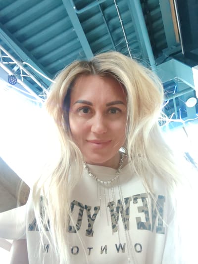 The media file shows a selfie of a woman with blonde hair, wearing a white hoodie with a leopard print design. She is wearing a silver necklace with multiple chains. The background appears to be indoors, with a blue ceiling structure and some lighting fixtures visible. The lighting is bright and appears to be coming from the right side of the image, creating some overexposure. The woman is smiling and looking at the camera. There is text on her hoodie that appears to read "LOY NEW" and "MOTW" in large, block letters. The overall impression is a casual, personal photograph.