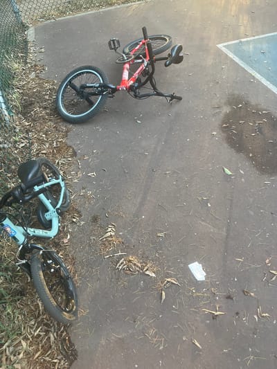 Two BMX bikes are lying on a paved surface near a chain-link fence. One bike is red and black, and the other is light blue and black. The red bike is upright, while the blue bike is on its side. The ground is littered with leaves and debris. The scene appears to be outdoors, possibly a park or a schoolyard, with a white painted line on the pavement suggesting a sports court. There is some dappled sunlight, indicating it is daytime. A small white piece of paper is visible on the ground between the two bikes. No people are present in the image. No text is clearly legible on the bikes or surroundings.