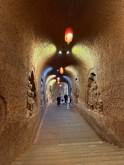 The image shows the Shizhong Temple in China. The location is a man-made tunnel carved out of the rock, with stone and wall carvings, some appearing damaged. Several people are walking through the tunnel in the distance. Hanging red lanterns are strung along the tunnel's ceiling, and the walls are textured and rough. There are stone slabs along the right side of the tunnel floor.