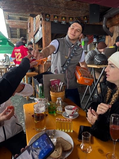 The image displays an outdoor bar area in St. Christoph, Austria, characterized by rustic wooden beam construction with snow and slush visible on the ground outside. Banners featuring "DIE NACHT MACHT ALLE" hang from overhead beams.
In the center, a male subject with short dark hair, wearing a black cap, sunglasses on his cap, a grey sleeveless vest with a Jägermeister logo, and a white t-shirt, extends a pair of orange plastic sunglasses towards a partially visible hand on the left. His left arm holds an orange box labeled "Jägermeister ORANGE."
Seated at a light brown wooden table in the foreground, a female subject on the right has long blonde braided hair and wears a white beanie and a black fur-trimmed jacket, with her hands clasped on the table. Visible items on the table include: a glass of beer, a glass of green liquid, a white teacup with a red design on a saucer (containing a red sugar packet and an empty creamer), a wooden caddy filled with various metal utensils, a green glass bottle of water, a clear glass bottle, a white plate with two slices of dark bread and a white substance, five small glass vials containing yellow liquid, and two stemmed glasses, one holding a reddish liquid and one empty. A dark smartphone is near the female's hands, and another smartphone displaying a lock screen with the time "12:44" and a mountain image is held in the lower-left by an unseen individual.
In the background, a person wearing a red jersey with "O'CONNOR 22" on the back is visible. To the right, another male subject wearing a brown beanie is seated at a separate table, with an "Oakley" ski goggle case placed beside him. A speaker is mounted on a wooden beam, and a green canopy is partially visible in the far left background.