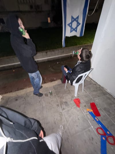 The scene depicts an outdoor gathering at night, likely on a patio or balcony, in Migdal HaEmeq, Israel. Three individuals are present. To the left, a person wearing a black hoodie and blue jeans stands, raising a green beverage can to their mouth. To the right, another person is seated on a white plastic chair, holding a green beverage can to their mouth with their right hand while smoking a cigarette held by their left hand. This individual wears a dark jacket. In the foreground, the lower torso and legs of a third individual are visible, wearing a dark zippered hoodie and light-colored pants. An Israeli flag with a blue Star of David on a white field, bordered by blue stripes, hangs vertically in the background. The ground is paved with rectangular concrete tiles. Adjacent to the tiled area, a low concrete curb separates it from a dark, unidentifiable ground area, possibly grass. Scattered on the tiled ground are an empty green beverage can, a piece of red fabric, and a large blue and red plastic toy resembling oversized scissors. The area is illuminated by artificial light, and the background beyond the curb shows blurred elements of what appear to be residential buildings or foliage under nighttime conditions.