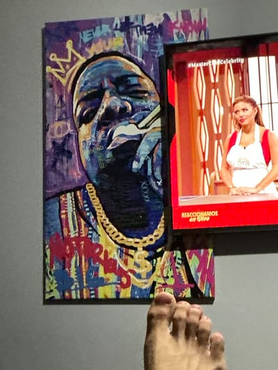 A television screen shows a celebrity chef on a red background with the hashtag #MasterChefCelebrity. Below the TV is an abstract painting on a rectangular board, depicting a face with a crown and gold chain. The artwork features vibrant colors and mixed textures. A person's toes are partially visible, positioned near the edge of the painting. The background is a plain gray wall.