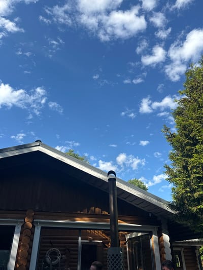 The image captures an outdoor scene in Saue, Estonia, under a bright, blue sky dotted with fluffy white clouds. The perspective is from below, looking upwards towards the sky and the side of a wooden building, possibly a sauna or cabin, with a dark roof.
Green foliage from a tree drapes over the top edge of the frame, suggesting the building is situated in a natural or semi-rural environment. A metal chimney pipe protrudes from the roof. The building's exterior is constructed from logs and features several windows. In one window, a white circular object with text is visible, reading "MINÄ MAAILMAN LINN..." which appears to be Finnish for "I the world's bird...". A person's head and shoulder are partially visible in the upper right corner of the frame, looking out of the window.
The lighting suggests it is daytime, likely late morning or afternoon, with sunlight casting shadows on the building. The overall atmosphere is peaceful and summery.