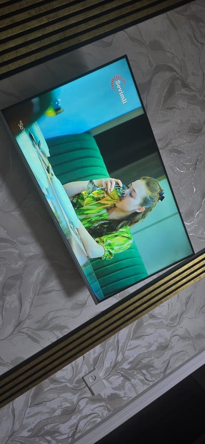 The image shows a television screen displaying a woman in a green outfit drinking from a glass. The television is mounted on a wall with textured wallpaper. There is a decorative element on the wall above the television, consisting of horizontal dark and gold stripes. Below the television, an electrical outlet is visible on the wallpaper. The scene is likely a living room or entertainment area. There is no identifiable information in the image to determine its location in the city of Uychi, Uzbekistan.