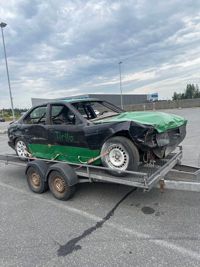 The image shows a heavily damaged black and green sedan on a trailer in Slitu, Norway. The car appears to be a demolition derby or race car, with its windows removed and a roll cage visible inside. The front of the car is severely crumpled, and the hood is painted green and is heavily dented. The side of the car has "Tiril" and a heart symbol spray-painted in green. The trailer is metallic grey with two axles and black fenders. The car is secured to the trailer with orange straps. The background shows a grey, cloudy sky, a light pole, a building with a blue sign that reads "BILTEM", and some trees. The scene is set in what appears to be a parking lot. There are no people visible in the image.