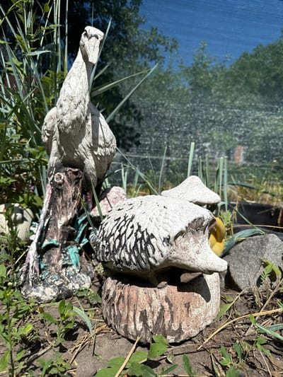 In Valky, Ukraine, this image depicts a garden scene featuring two concrete garden ornaments. The primary subject on the left is a tall, white bird statue, possibly a heron or crane, standing on a weathered, brown log. The log has green and pink paint accents. To the right of the bird statue, a smaller, white hedgehog statue is positioned on a rounded, reddish-brown base that resembles a tree stump. The hedgehog has a textured surface with black markings that mimic quills. Both statues appear to be aged and weathered. The garden is overgrown with tall green grass and other plants, some of which are in focus and some are blurred. In the background, a blue net screen is visible, likely serving as a shade or privacy barrier, with the blurred shapes of trees and a bright sky visible through it. The ground is covered in dark soil, dry grass, and a few small green plants. The overall impression is one of a slightly neglected, yet characterful, outdoor space.