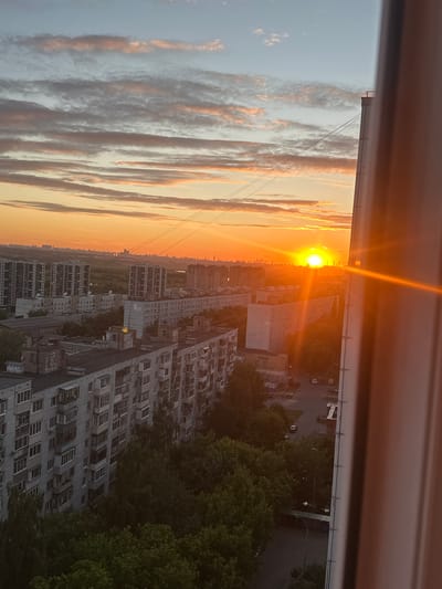 The image is a vertical, high-angle shot capturing a sunset over a densely populated urban area, likely Lytkarino, Russia. The sky is a gradient of soft blues and oranges, with wispy clouds illuminated by the setting sun, which is prominently featured in the middle right of the frame, casting a bright orange glare. In the foreground, on the right, a portion of a light-colored building or structure obscures part of the scene, with a warm orange glow reflecting off its surface.
The mid-ground is dominated by numerous multi-story apartment buildings of varying architectural styles, many appearing to be Soviet-era panel buildings, characterized by their uniform facades and numerous balconies. These buildings are packed closely together. Between the buildings, pockets of green trees are visible, suggesting courtyards or small parks. Power lines crisscross the sky above the buildings.
The background shows a hazy cityscape with more buildings, stretching towards the horizon where the sun is setting. The overall impression is a residential district bathed in the warm, atmospheric light of sunset. No individuals are clearly discernible in the image. The scene is static, devoid of overt action, and appears to be a typical view from an elevated apartment window.
