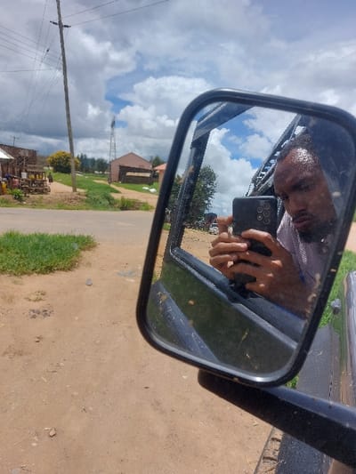 The image captures a scene from a stationary vehicle on an unpaved road in Kimamba, Tanzania. A black-framed side mirror occupies the central-right portion of the frame, reflecting a male individual taking a selfie. The man, visible from the shoulders up, has short dark hair and a beard, with visible perspiration on his forehead, and is wearing a light-colored, possibly striped, collared shirt. He is holding a dark-colored smartphone with a multi-lens camera module directly towards his reflection.
The external environment, viewed around the mirror, features a reddish-brown dirt road with patches of green grass on the left. In the mid-ground, a tall wooden utility pole with power lines is positioned on the left. Further back, what appears to be a small wooden market stall with goods displayed on a table is visible. In the background, a light-colored building with a darker roof section is present, behind which a tall metal lattice tower (possibly a cell or water tower) extends into the sky. The sky is partly cloudy with significant white cloud formations and patches of blue. The vehicle's dark exterior bodywork below the mirror shows visible dirt accumulation.