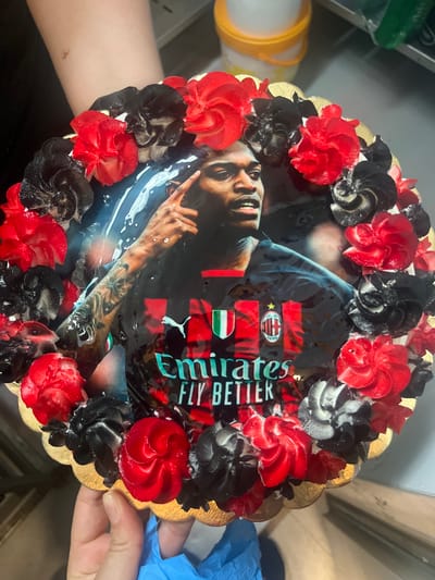 This media file is an overhead shot of a round cake decorated with red and black frosting swirls, resembling flowers. The cake is placed on a golden scalloped cake board and is being held by two hands, one wearing a blue glove. The cake's surface features a glossy edible image of a man with tattoos on his arm, wearing a dark shirt with a soccer team logo and text that appears to say "Emirates FLY BETTER." The image has a celebratory feel, likely for a fan of the soccer team depicted. The background is blurred, but suggests an indoor setting with some kitchen-related objects visible, including a yellow container. The lighting is bright and even, suggesting it was taken during the daytime. No specific time of day, weather, or strong emotions are discernible beyond the celebratory nature of the cake. The location context provided is Vallo della Lucania, Italy.