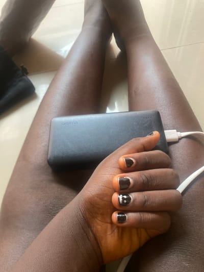 A close-up shot shows a person's hand resting on a dark gray power bank, which is plugged into a white cable. The hand has dark skin and is adorned with black nail polish. One nail on the index finger is painted with a simple smiley face. The power bank has the "oraimo" logo printed on its side. The scene appears to be indoors, on a tiled floor, with the lighting suggesting daytime. The person's legs are also visible, with one leg bent and resting on the other. The overall impression is casual and relaxed, possibly indicating the person is charging their device while taking a break. The location context is Wauro Leggal, Nigeria.