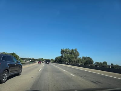 The image depicts a view from inside a vehicle traveling on a multi-lane highway under a clear blue sky. The highway appears to be in a suburban or rural area with trees and vegetation lining the right side. On the left, there is a dark-colored SUV in the foreground, and further ahead, other vehicles are visible, including a red sedan and a black utility vehicle. An overpass or bridge structure can be seen in the distance. The road surface shows lane markings and some wear. The overall scene suggests a sunny day with moderate traffic on a highway. Based on the provided information, this scene is located in West Sacramento, USA.