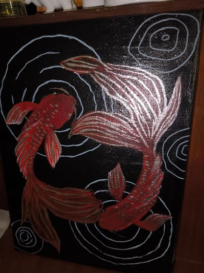 The image displays a black canvas painting of two red koi fish swimming in a circular pattern, symbolizing yin and yang. The fish have metallic silver accents on their fins and tails, and the water is depicted with white swirling lines. The painting is framed by a wooden border. The setting is indoors, with a shelf above the painting holding various objects, including a glass jar and a white figurine. The overall scene appears to be a domestic interior. There is no indication of the city of Bolshaya Martynovka, Russia, in the provided image or its context.