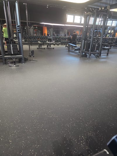 A brightly lit gym with weight machines and equipment. The location is likely Zirndorf, Germany, though not verifiable from the image. There is one individual using a weight machine and a few others in the distance. The floor is dark with light-colored speckles. There are windows allowing light into the facility.