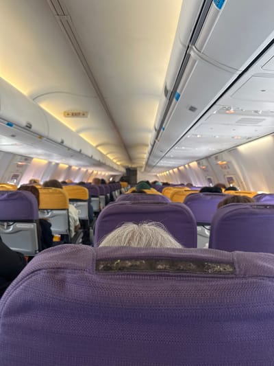 The image shows the interior of an airplane cabin, likely at Phuket International Airport in Mai Khao, Thailand. Rows of purple and yellow airplane seats are visible, occupied by passengers. Overhead compartments and lighting fixtures line the ceiling of the cabin. An "EXIT" sign is illuminated above the aisle. The perspective is from a passenger seated in the back of the plane, looking forward. The overall lighting suggests an artificial light source within the cabin.