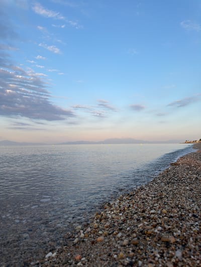 This is a low-angle, full shot of a pebble beach meeting the calm sea under a pale blue sky with scattered clouds. The shore is covered in smooth, multi-colored pebbles ranging in size from small grains to about two centimeters in diameter. The sea is a clear, light blue with gentle ripples. The horizon line is marked by distant, hazy mountains and a faint line of buildings along the coast, suggesting the location is near a town. The sky transitions from a brighter blue overhead to a softer pinkish hue near the horizon. There are no people or animals visible in the image. The scene appears to be a serene coastal landscape in Νέα Φλογητά, Greece, possibly at dawn or dusk.