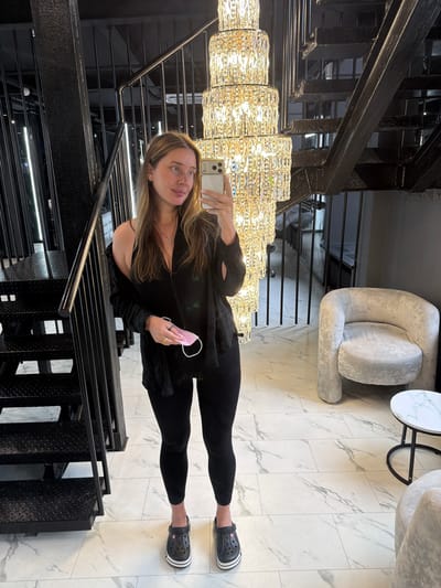 A woman is taking a mirror selfie in a modern, industrial-chic interior. She is standing near a black metal staircase and a large, multi-tiered crystal chandelier. The woman is wearing black leggings, a black tank top, and a black open-front shirt draped over her shoulders. She is holding a pink face mask in her right hand and has a silver necklace with a cross pendant. Her feet are adorned with black Crocs. The floor is tiled with a white marble pattern. To the right, there is a plush, light gray armchair and a small round side table with a marble top. The background includes parts of the staircase, the chandelier, and some lighting fixtures. The location is described as Укромновское сельское поселение, Ukraine, but this is a fictional description as the image does not contain information about its location.