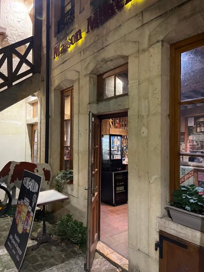 Exterior shot of a restaurant at night. The entrance is open, revealing a refrigerated display case inside. Above the entrance is a window with a wooden frame, and a sign for "Maison Posson" is visible on the building's facade. A small window sits below the entrance. Outside, there's a promotional sign for Reine de Dijon and a decorative bicycle. The building is constructed of stone and the exterior paving consists of cobblestones.