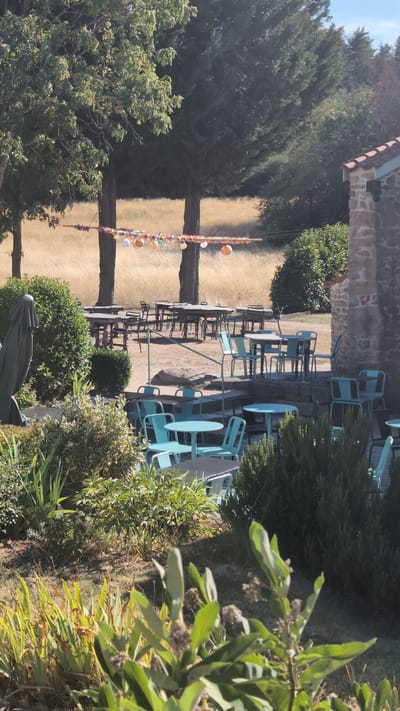 The image depicts an outdoor seating area of a restaurant or cafe in Chambles, France. The scene is set against a backdrop of trees and a dry, golden field, suggesting a warm climate or season. In the foreground, there is lush green vegetation and foliage.
The main focus is on the arrangement of tables and chairs. Several round and square tables, some with dark tops and others in a light turquoise color, are placed outdoors. These are accompanied by numerous chairs, also predominantly in a light turquoise hue, as well as some dark metal chairs. The furniture is scattered across a gravelly or dirt ground, with some tables and chairs situated on stone steps.
In the middle ground, more tables and chairs are visible, appearing to be set up for dining. Behind this seating area, two large trees stand prominently, with a string of colorful, small lanterns or decorations strung between them. Beyond the trees, the expansive field of dry grass stretches out, bathed in sunlight.
To the far right, a portion of a stone building with a tiled roof is visible, indicating the establishment's architecture. A dark, draped statue or figure is partially visible on the left side of the image, near some dense green bushes. The sky is clear and blue, with bright sunlight casting shadows and highlighting the textures of the scene.