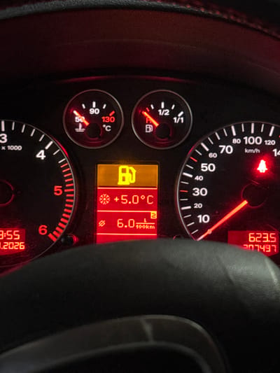 A close-up, front-facing image captures the illuminated instrument cluster of a vehicle, located in Radomirtsi, Bulgaria. The dashboard is primarily black with red backlighting for dials and needles. On the left, a partially visible tachometer is present. Above the digital display, an analog coolant temperature gauge indicates a temperature just below 90°C, and an analog fuel gauge shows the fuel level as critically low, almost at the empty mark.
The central digital display panel is divided into three sections:
1. Top: A prominent yellow warning light depicts a fuel pump icon, signaling low fuel.
2. Middle: Displays the external temperature as +5.0°C, indicated by a snowflake symbol.
3. Bottom: Shows an average fuel consumption of 6.0 L/100km.
To the right of the central display, an analog speedometer reads approximately 0 km/h, indicating the vehicle is stationary or moving very slowly. Below the 10 km/h mark, a red warning light depicting a buckled person (seatbelt reminder) is illuminated. Digital readouts at the bottom-left of the cluster show the time as 9:55 and the date as 1.2026 (January 2026). At the bottom-right, a trip mileage of 623.5 is displayed above a total odometer reading of 207497 kilometers. A segment of a dark, textured steering wheel rim is visible in the foreground, partially obscuring the lower edge of the instrument cluster.