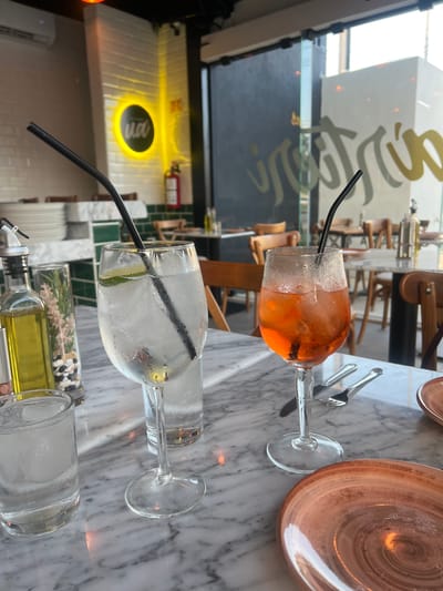 This media file captures a close-up, eye-level view of a dining table setting at what appears to be a modern restaurant or cafe in Mérida, Mexico, during the daytime.

**Subjects:** The primary subjects are two beverages served in stemmed glasses on a white marble table. One glass holds an orange-hued drink with ice and a black straw, resembling an Aperol Spritz. The other contains a clear drink with ice, a lime wedge, and a black straw, likely water or a clear spirit. Also on the table are two brown ceramic plates, cutlery (a fork and knife), and a glass bottle containing oil and herbs (rosemary and possibly small decorative stones), along with another glass that could be for ice or another drink.

**Scene and Setting:** The background reveals an indoor dining area with wooden chairs and tables. A large window reflects the bright outdoor environment and some text. Further inside, there's a wall with a prominent circular yellow glowing sign, possibly part of the establishment's branding.

**Activity or Event:** The arrangement of drinks, plates, and cutlery suggests a casual meal or a relaxed drink break at a restaurant.

**Notable Details:** The glasses exhibit condensation, indicating the drinks are cold. Bright natural light streams in through a large window, suggesting it is daytime with potentially clear weather outside. The overall aesthetic is clean and modern, with the marble table and simple glassware.

**Visible Text:** Reflected on the window glass in a script font, the word "Solera" or a similar variant starting with 'S' is partially visible. In the background, a circular yellow sign features a dark center with white text or a logo, appearing to read "100%" or "100".