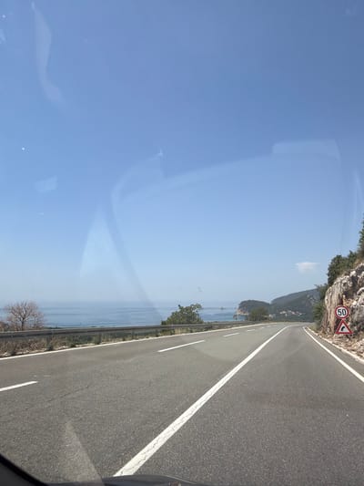 The image depicts a scenic coastal road in Kanjoši, Montenegro, on a clear, sunny day. The perspective is from the driver's seat of a vehicle, looking forward along the asphalt road. The road is divided by white lines and curves gently to the right. To the left of the road, there is a metal guardrail bordering a steep drop-off. Beyond the guardrail, the Adriatic Sea stretches to the horizon, with a few small islands visible in the distance. The right side of the road is bordered by a rocky cliff face covered in vegetation. A speed limit sign indicating "50" and a warning sign for a curve are visible on the right side of the road. The sky is a clear, bright blue. The overall impression is one of a beautiful, winding road through a picturesque coastal landscape.