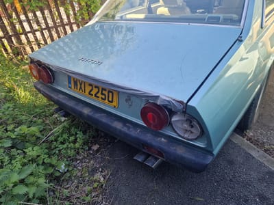 This media file depicts the rear-right quarter of a light blue/green vintage car, likely a Ferrari given the emblem on the trunk lid. The car appears to be in a state of disrepair, with noticeable damage to the rear-right taillight assembly, where the outer lens is missing, exposing the bulb and interior reflector. The matching left taillight remains intact.
Visible on the trunk lid is the embossed text "FERRARI". A yellow British number plate, displaying "GB" and the registration "WXI 2250", is centrally located on the rear. Below the bumper, two metal exhaust pipes are visible. Peeling trim and some rust are apparent along the edge of the trunk lid and bumper. Through the car's rear window, parts of the interior, including beige seating and a "PIONEER" branded object, can be seen. An antenna is mounted on the rear-right pillar.
The car is parked outdoors during daylight hours, with clear natural light illuminating the scene. It is situated adjacent to an overgrown area of green grass and weeds, bordered by a rustic wooden fence in the background. The car's right side is on a dark grey paved surface, likely tarmac or asphalt, next to a concrete curb. The overall impression is that of a classic vehicle that has been neglected or abandoned, possibly in a garden or a quiet side area in South Tyneside, United Kingdom. No people or active events are present in the image.