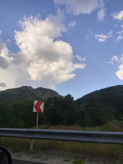 The image shows a landscape with a mountain range, a blue sky with white clouds, and a road with a guardrail and a road sign indicating a sharp turn to the left. The mountains are covered with green vegetation. The photo was likely taken from a moving vehicle. The exact location in Bukovo, Bulgaria, cannot be determined from the image.