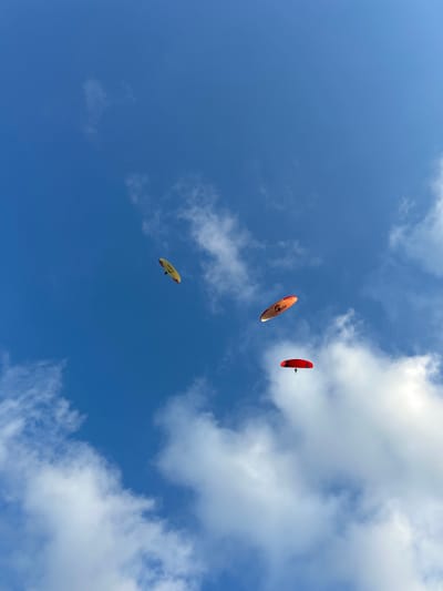 In the blue sky above Mekar Sari, Indonesia, three paragliders are visible against a backdrop of scattered white clouds. The paragliders are at different altitudes and positions within the frame. The topmost paraglider is yellow and appears to be higher and further to the left. Below and to the right of the yellow paraglider, an orange and white paraglider is seen, with text that appears to read "Skydiving" on its wing. The lowest paraglider is a bright red, positioned in the lower right quadrant of the image, with a person suspended below its wing. All three paragliders are in flight, suggesting an activity taking place in an open area, likely outdoors given the sky and clouds. The image is captured from a low angle, looking directly up at the paragliders and the sky.