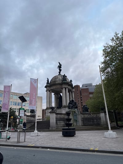 This image depicts the Queen Victoria Monument in Liverpool, United Kingdom. The monument is a large, ornate structure made of stone with columns and sculptures. A winged figure stands atop the dome.
The monument is situated on a paved area alongside a road. The sky is overcast with grey clouds.
There are several banners that say "Stay for Art." A building with windows is visible in the background. The architecture of nearby structures indicates a city environment.