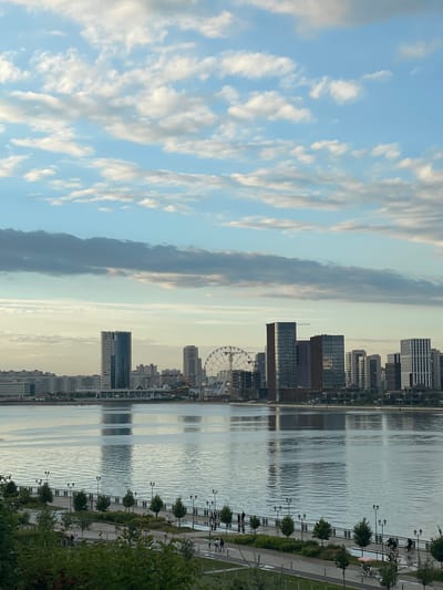 The image depicts a waterfront city view with a prominent Ferris wheel and modern skyscrapers. The scene is set during the day, under a sky with scattered white and gray clouds. In the foreground, a paved walkway with lampposts and trees lines a calm body of water. Several individuals are visible walking and cycling along the path. Across the water, a cityscape features a variety of buildings, including tall, glass-fronted structures and lower-rise residential buildings. The water reflects the sky and the buildings, creating a shimmering effect. The location is identified as Дачное, Russia.