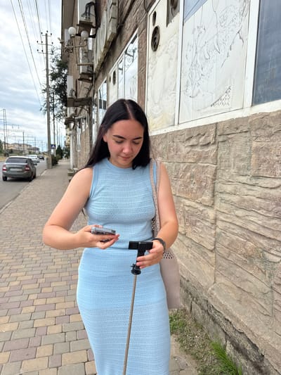 A young woman with dark hair is taking a selfie with a smartphone attached to a selfie stick. She is wearing a light blue knitted dress and a beige bag is slung over her shoulder. She is standing on a cobblestone sidewalk next to a building with a stone facade and large windows that reflect the surroundings. The woman has a slight smile on her face. In the background, there is a street with parked cars and utility poles with wires. The scene appears to be outdoors in daylight, with an overcast sky. The presence of cobblestone streets and the architectural style of the building suggest an urban or historic setting. The text "Derbent, Russia" is provided as location context.
