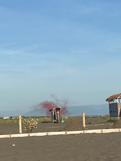 The image depicts an outdoor scene with a clear blue sky. In the mid-ground, there is a small, dark wooden structure with a pitched roof, resembling a shed or a toilet. Red smoke is emanating from behind and above this structure, suggesting it might be a signal or a deliberate emission. To the right of the shed, a larger structure with a thatched roof is partially visible, appearing to be a covered seating area or a kiosk. In the foreground, a dark sandy or dirt surface stretches across the frame, with a white concrete curb running along the bottom edge. A thin, leafless shrub grows near a wooden post in the mid-ground. In the background, distant mountains or hills are visible under a hazy sky. The overall environment appears to be a coastal or open area, possibly a beach or a park. There are no identifiable people or animals in the scene. The location is identified as the city of Pistula, Montenegro.