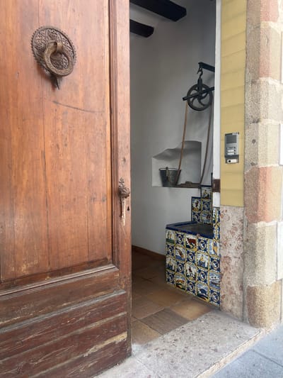 The image captures an entryway featuring a large, weathered wooden door that is ajar, offering a view into an old building's interior. The door itself is made of wooden planks, showing signs of age and wear. A prominent, ornate circular metal door knocker or handle with intricate patterns is affixed to the door.
Through the opening, a section of a white-walled interior is visible, dominated by a rustic well or water retrieval system. This includes a robust cast-iron pulley mounted on a bracket, with a thick rope extending downwards into a cutout in the wall where a metal bucket is positioned. Dark wooden beams are also visible within the interior.
Above the door and extending across the archway, the structure is composed of rough-hewn stone blocks in varying shades of grey and reddish-brown. A band of light yellow rectangular tiles runs beneath these stones. Mounted on these yellow tiles, to the right of the door's opening, is a grey intercom unit with visible buttons and a speaker grill. The brand name "FERMAX" is faintly discernible on this unit.
To the right of the door and below the yellow tiles, a wall section is decorated with distinctive blue, white, and yellow patterned ceramic tiles (azulejos), depicting various figures and geometric designs. The ground immediately outside the door features plain brown square tiles, transitioning to a textured grey stone surface further right. The scene conveys a sense of history and traditional architecture, consistent with its given location context of Sant Pol De Mar, Spain, and appears to be captured during the daytime.