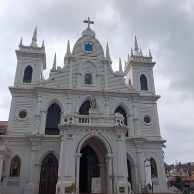 The image shows the Milagres Church in Marna, India. It is a large white church with a cross on top, various architectural details including arched windows and doorways, spires, and statues. The sky is overcast. There are also signs posted near the entrance of the church.