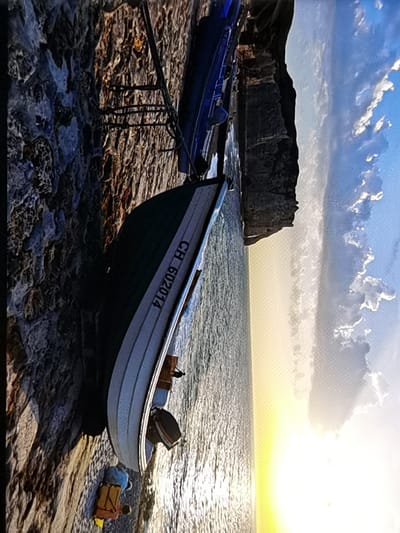 The image displays a tranquil scene at the base of the cliffs of Étretat, France. A small boat, identified by the markings "CH 602014," rests on the rocky shoreline. The boat is partially submerged in the calm, shimmering water, suggesting it has been pulled ashore or is waiting for the tide. Beside the boat, there are two bags, one blue and one yellow, suggesting a recent presence or belongings left behind. In the background, the iconic white cliffs of Étretat rise majestically, with a clear blue sky dotted with soft clouds overhead. The sunlight, low in the sky, casts a warm glow over the scene, creating a serene and picturesque atmosphere characteristic of this renowned coastal location.