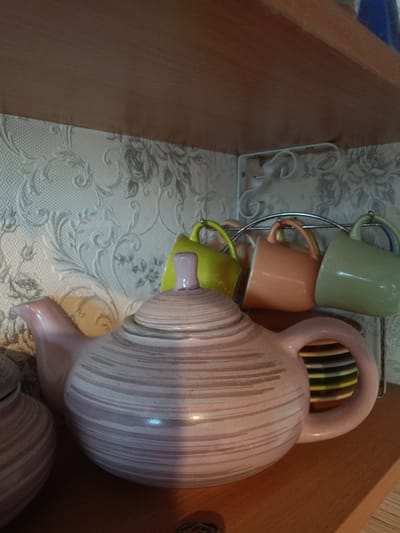 A close-up shot captures a collection of ceramic tableware items resting on a wooden shelf. The foreground is dominated by a large, light pink teapot with subtle, horizontally painted stripes. To the left of the teapot, a portion of another, smaller ceramic container with similar striped detailing is visible. Behind the teapot, a metal rack holds four colorful mugs: one lime green, one peach, and two yellow. These mugs are suspended by their handles. The background features a textured wallpaper with a floral pattern in shades of white and gray. The scene appears to be an indoor domestic setting, possibly a kitchen or dining area. There is no discernible information to connect this image to Petrozavodsk, Russia.