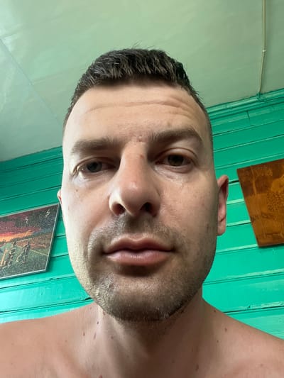 A close-up, low-angle selfie captures a man's face, looking directly at the camera. He has short, dark hair, a light complexion, and stubble on his chin and jawline. His eyes are open, and his lips are closed in a neutral expression. The background consists of a teal-colored wall made of horizontal planks. To the left of the man's head, a framed picture with a landscape scene is visible. To the right, another framed object, possibly a piece of art or decoration, with a darker, textured surface is present. The lighting suggests an indoor setting, possibly during the day. The image does not provide any specific landmarks or clues to pinpoint its location within Navahrudak, Belarus.